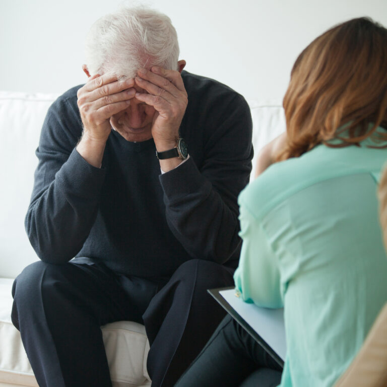 Un homme d'âge mûr, assis dans le cabinet d'un psychiatre, se tenant la tête dans les mains en signe de détresse. En face de lui, un professionnel de santé mentale prend des notes, symbolisant l'écoute et le soutien psychologique.