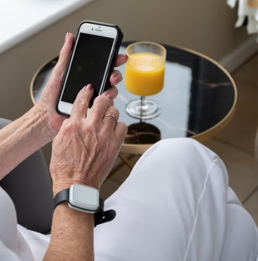 Une femme assise dans un fauteuil consulte son téléphone en toute sérénité grâce au bracelet anti-chute qu'elle porte dans le cadre de son pack de téléassistance